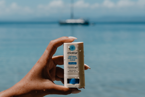 Hand holding a zinc sunscreen stick with the ocean and a boat in the background