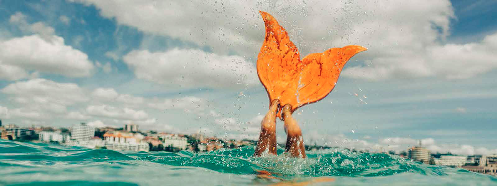Person in orange mermaid tail splashes in ocean with city skyline and bright, cloudy sky.